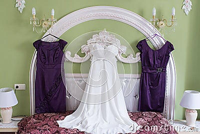 White wedding dress and two lilac bridesmaidâ€™s dresses on a hanger on the wall. Morning of the bride. Fees. Stock Photo